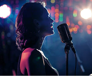 Person singing into a microphone with multicolored stage lights in background.